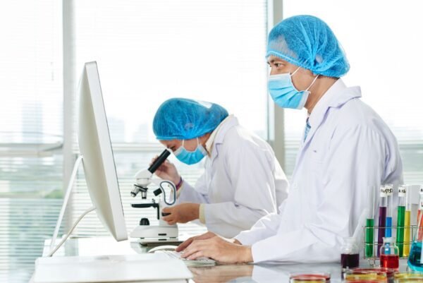 Team of talented microbiologists wearing white coats and medical masks wrapped up in work at modern lab: Asian woman studying sample with help of microscope while her male colleague taking notes